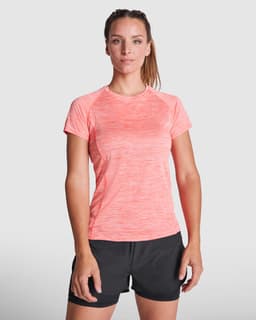 A woman stands in a coral pink sports T-shirt paired with black shorts, featuring a simple logo on the shirt.