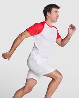 A man in a red and white T-shirt runs, wearing white shorts. The shirt features a logo on the front.