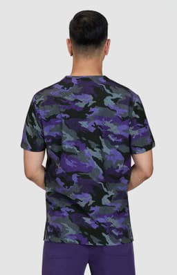 A camouflage print t-shirt in shades of purple, green, and black, viewed from the back. The model is wearing purple pants.