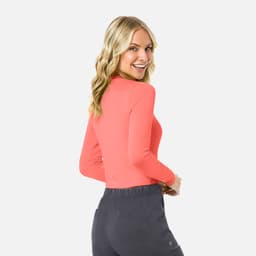 A woman models a long-sleeve, coral underscrub tee, paired with dark grey pants. The shirt features a branding logo.