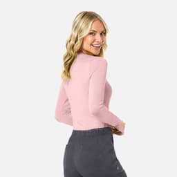A woman wearing a fitted pink underscrub tee and grey pants, smiling and facing slightly backwards.