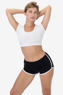 A model poses in black interlock running shorts with white trim, paired with a white crop top. The image features a branding logo.