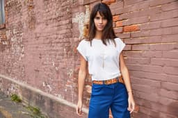 A light-coloured casual T-shirt, styled with blue trousers, worn by a model against a textured brick wall.