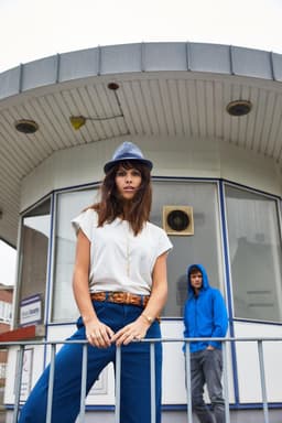 A woman in a white casual T-shirt and blue pants poses in front of a building, wearing a grey hat. A person in a blue hoodie is in the background.