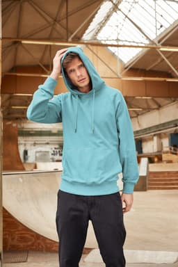 A teal men's hoodie featuring a front pocket and drawstring hood, worn by a model in an indoor skatepark.