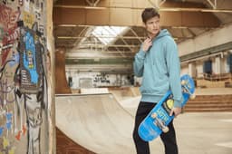 A male model in a blue hoodie holds a skateboard in a skate park setting, with graffiti on the walls.