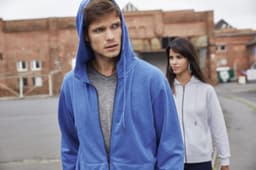 A blue zip hoodie and a grey zip hoodie worn by a man and a woman in an outdoor urban setting. Both hoodies have logos.