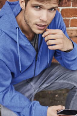 A blue men's zip hoodie is worn by a man sitting against a brick wall, holding a smartphone.