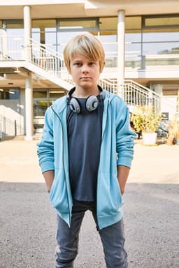A child in a light blue zip hoodie with headphones, standing confidently outdoors in a casual setting.
