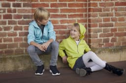 A blue zip hoodie and a lime green zip hoodie worn by a boy and a girl seated against a brick wall.