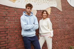 A light blue men's lounge hoodie and a cream women's hoodie are worn by two models against a brick wall backdrop.