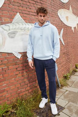 A model wearing a light blue men's lounge hoodie paired with navy pants, standing against a brick wall.
