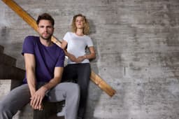 A purple T-shirt and a white T-shirt displayed by a man sitting on stairs and a woman standing nearby, both in casual attire.