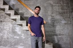 A men's purple T-shirt is displayed on a model standing next to a concrete staircase in a rustic setting.