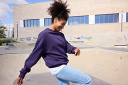 A purple oversized sweatshirt with a relaxed fit, worn by a person smiling near a skate park.
