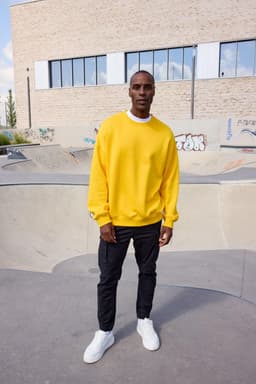 A person wearing an oversized yellow sweatshirt and black trousers stands in an outdoor skate park.
