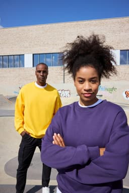 A purple oversized sweatshirt and a yellow oversized sweatshirt on two models against a textured wall backdrop.