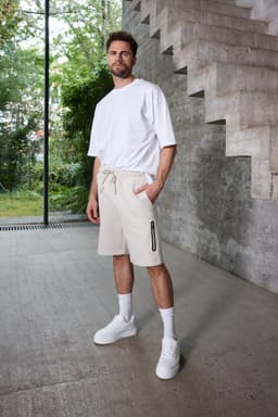 A man stands on a concrete surface wearing beige sports shorts and a white t-shirt, with a logo visible on the shorts.