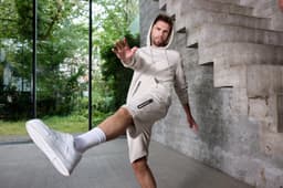 A man in light beige sports shorts and a matching hoodie is performing a kicking motion in a modern interior.