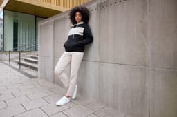 A woman in a colour-block black and cream hoodie leans against a grey wall, wearing beige trousers and white shoes.