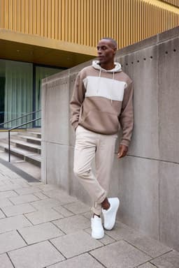 A colour-blocked sweat hoodie in earthy tones, paired with light beige pants and white sneakers, featuring a logo.