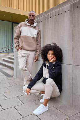 A colour-block hoodie in beige and black worn by two models, standing against a concrete wall, with casual pants.