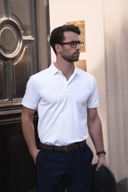 A man in a white polo shirt stands casually with his hands in his pockets, wearing dark trousers and glasses.