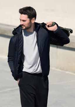 A man wearing a navy jacket with a hood, carrying a drink bottle over his shoulder. The background is a blurred outdoor setting.