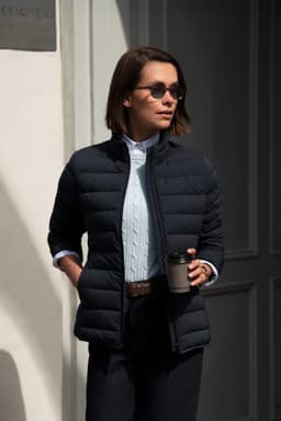 A woman wearing a navy blue down jacket, sunglasses, and holding a coffee cup, standing against a light background.