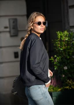 A woman wearing a black puffer jacket and sunglasses, walking past green foliage.