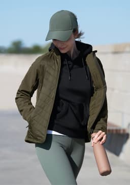 A woman wearing an olive green soft shell jacket over a black hoodie, paired with a cap and holding a drink bottle.
