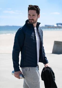 A man wearing a navy soft shell jacket, holding a drink bottle, with a beach and ocean in the background.