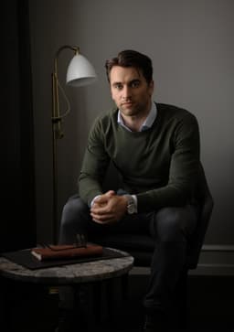 A man sits casually in a chair wearing a green Brighton O-Neck knitwear sweater, with a stylish lamp and table nearby.