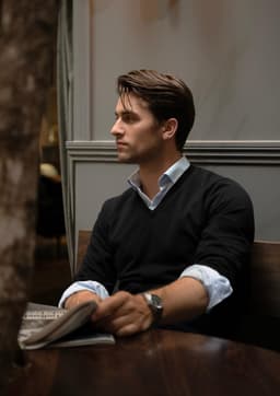 A man wearing a black V-neck jumper sits at a wooden table, holding a newspaper.