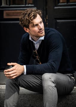 A seated man wearing a navy Brighton V-Neck knit sweater and grey trousers, holding a cup. The image features a logo.