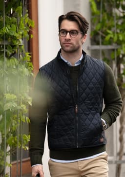 A man in glasses wears a navy quilted vest over a green sweater, walking outdoors with greenery in the background.
