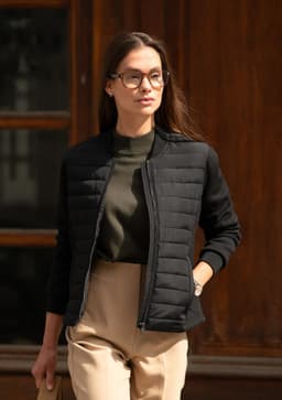 A woman in a black fitted jacket and olive green top poses outdoors, wearing glasses with light brown trousers.