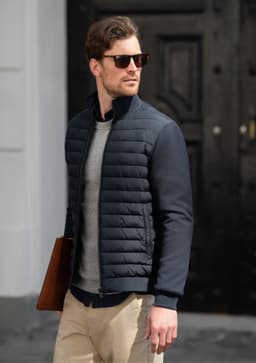 A man wearing a navy quilted jacket and sunglasses, holding a brown leather bag, walking in an urban setting.