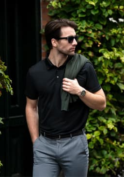 A black polo shirt paired with grey trousers, featuring a logo and styled with a green sweater draped over the shoulder.