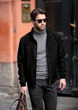 A man in a black jacket and grey turtleneck walks outdoors, carrying a brown bag. The jacket features a logo.