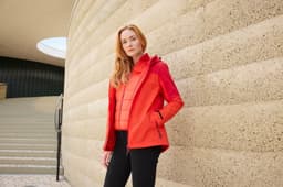 A woman wearing a vibrant red 3-in-1 jacket, standing against a textured stone wall, with a staircase in the background.