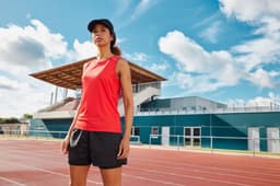 A woman in a red running tank top stands on an athletic track, with a sports facility in the background.