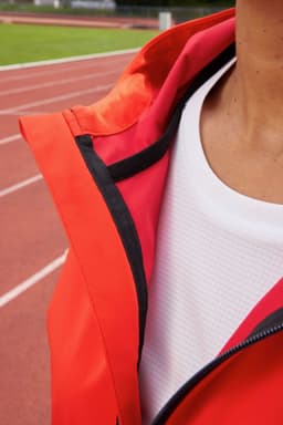 A close-up of a women's sports softshell jacket in vibrant red, featuring a hood and a black zipper.