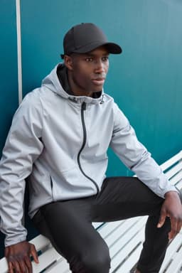 A grey men's sports softshell jacket with a hood, worn by a model seated on a bench, paired with a black cap.