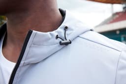 A softshell jacket in light grey, featuring a drawstring hood and a black zipper detail. It has a logo on the fabric.