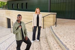 A unisex shirt jacket in olive green and beige worn by models on stone steps, featuring a logo.
