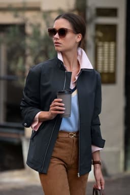 A woman is wearing a dark jacket with a light pink collar, holding a coffee cup in an outdoor setting.