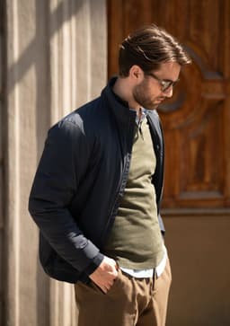 A man wearing a navy jacket over an olive sweater, standing outside next to a wooden door. The jacket has a logo.