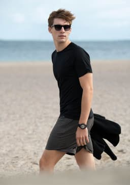 A young man wearing a black T-shirt and grey shorts walks on a beach, with sunglasses and a watch.
