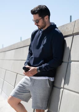 A navy hoodie with a branded logo, worn by a man leaning against a wall, paired with grey shorts.